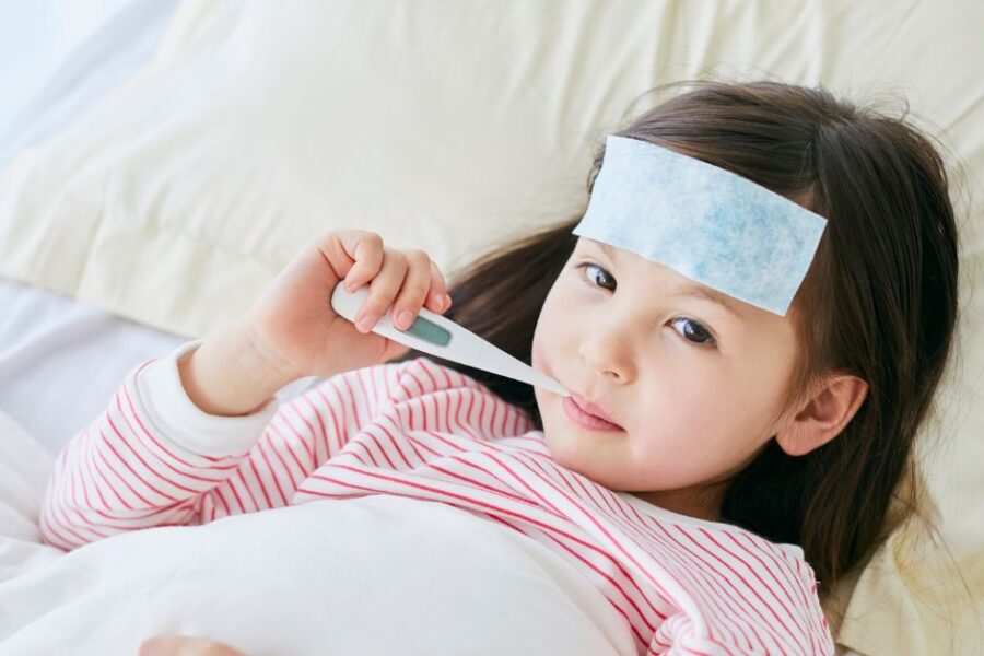 A young girl lays in bed with a thermometer in her mouth.