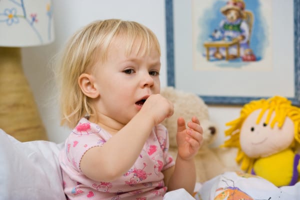 A fussy toddler sits in bed with her doll and teddy bear.