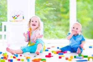 An adorable toddler girl and a baby boy play with colorful blocks while sitting on a floor in a sunny bedroom.