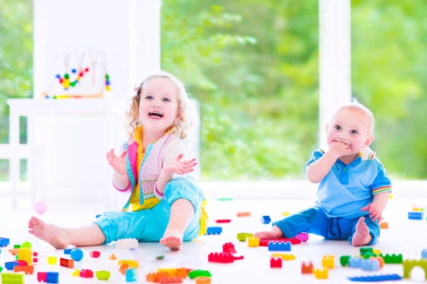 Adorable laughing toddler girl and a funny little baby boy, brother and sister, playing with colorful blocks sitting on a floor in a sunny bedroom with a big window