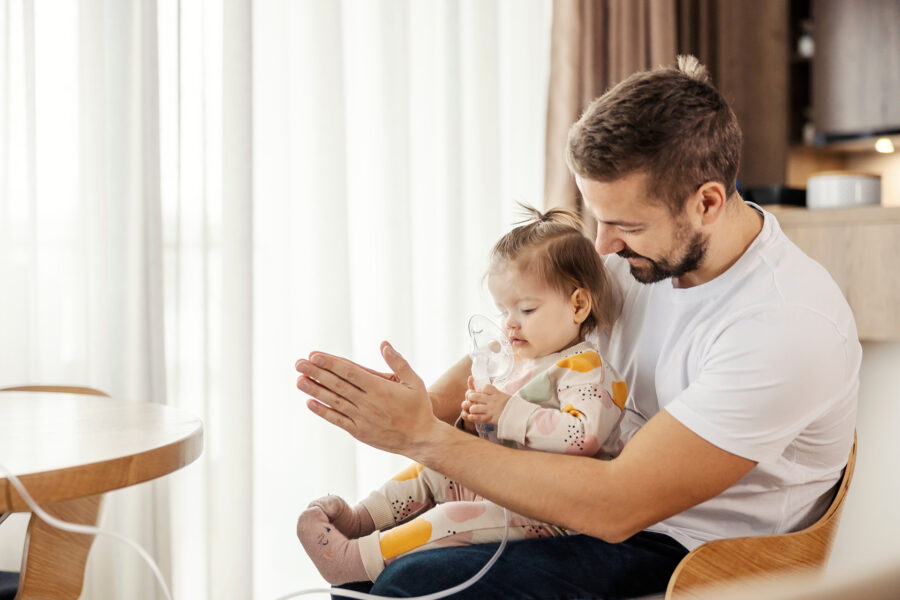 Father is holding his daughter in his lap while she using nebulizer.