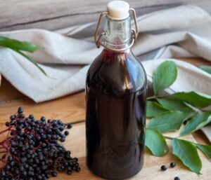 A bottle of elderberry syrup lays on a wooden surface with elderberries and a sprig of green leaves around it.