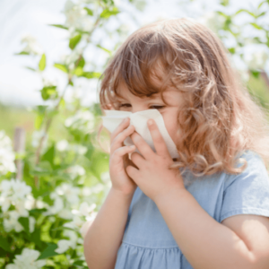 A young girl blows her nose while standing near a green bush with white blossoms. 