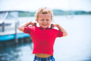 A young boy flexes his strong muscles as he plays near a lake. 