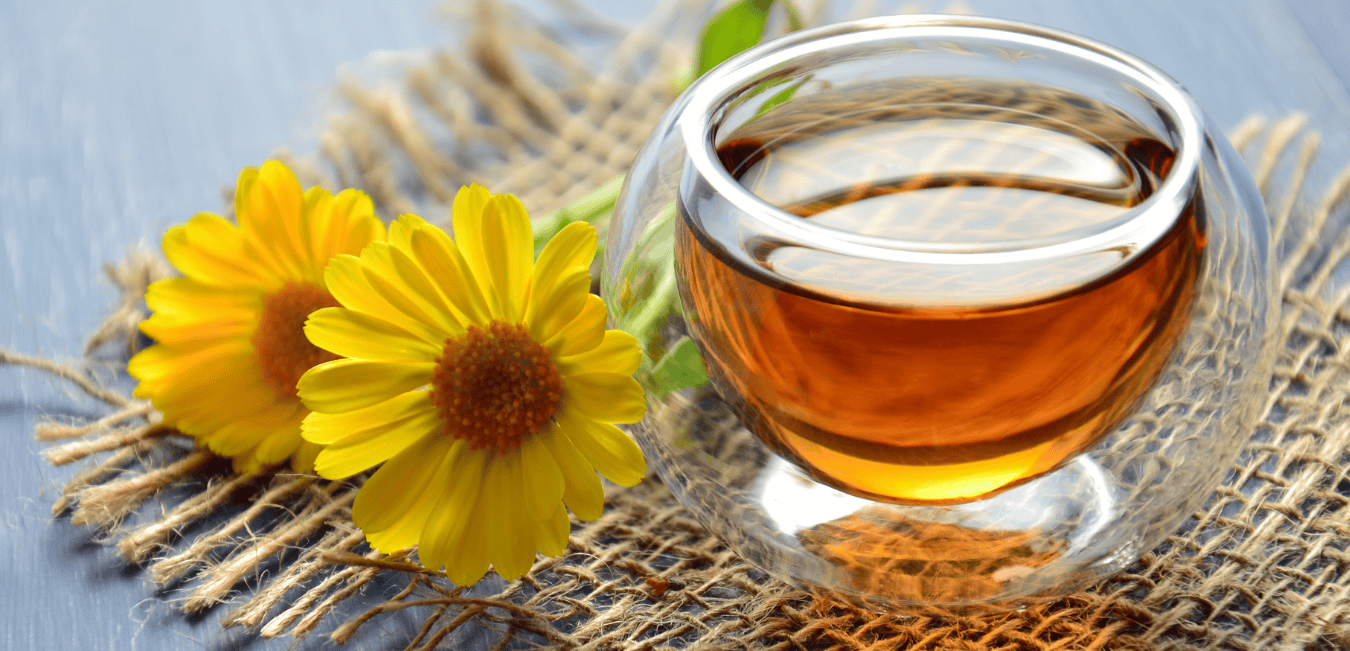 A cup of herbal tea sitting on a brown place mat next to a cheerful yellow flower.