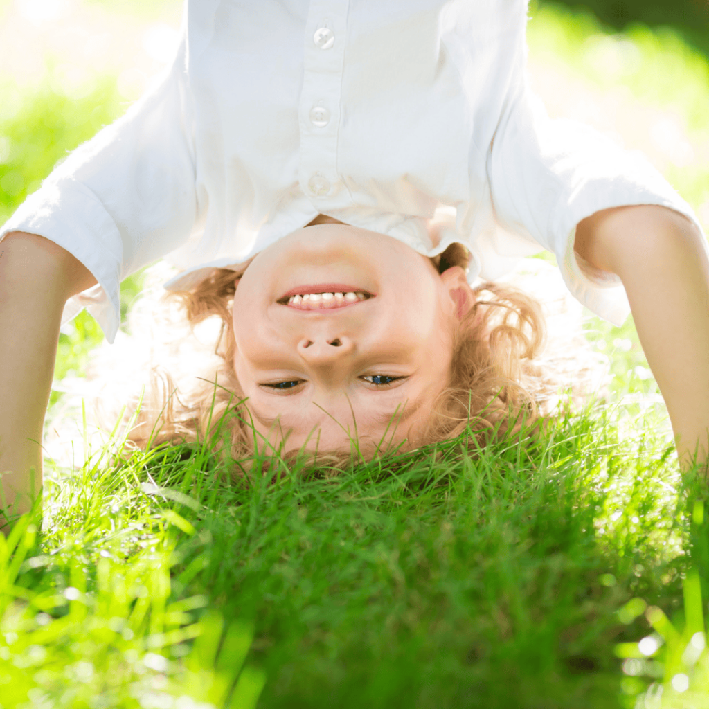 A smiling boy does a headstand in a yard with lush green grass. 