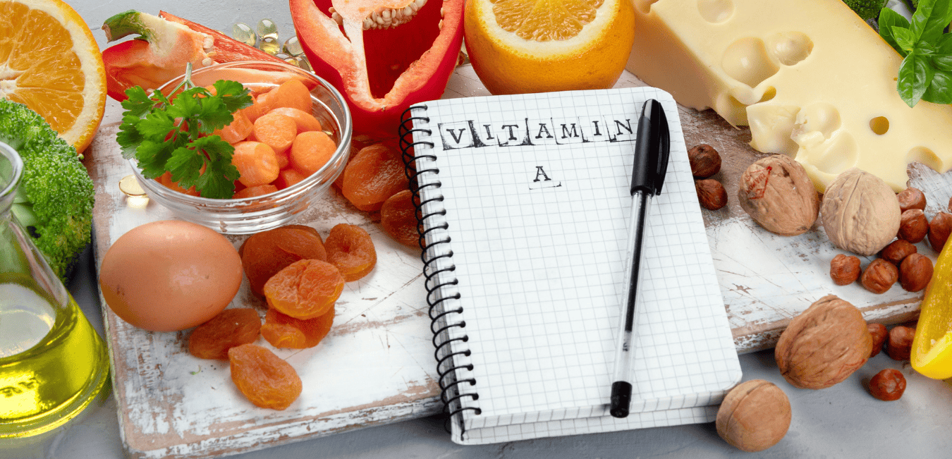 An open notebook with "Vitamin A" written on the page. Oranges, red bell peppers, and carrots surround the notebook.