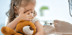 A young girl holding her teddy bear covers her eyes while receiving a shot in the arm.
