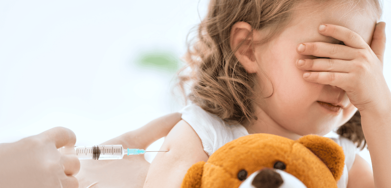 A vaccine is administered into the upper arm of a young girl holding her teddy bear.
