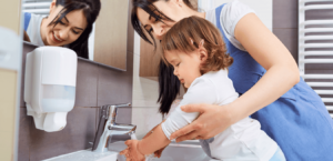 A mom helps her young child wash her hands in a bathroom sink.