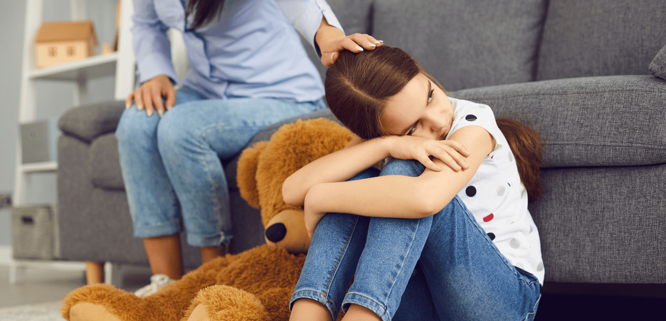 A distraught girl sits on the floor with her teddy bear while her mom gently strokes her hair.