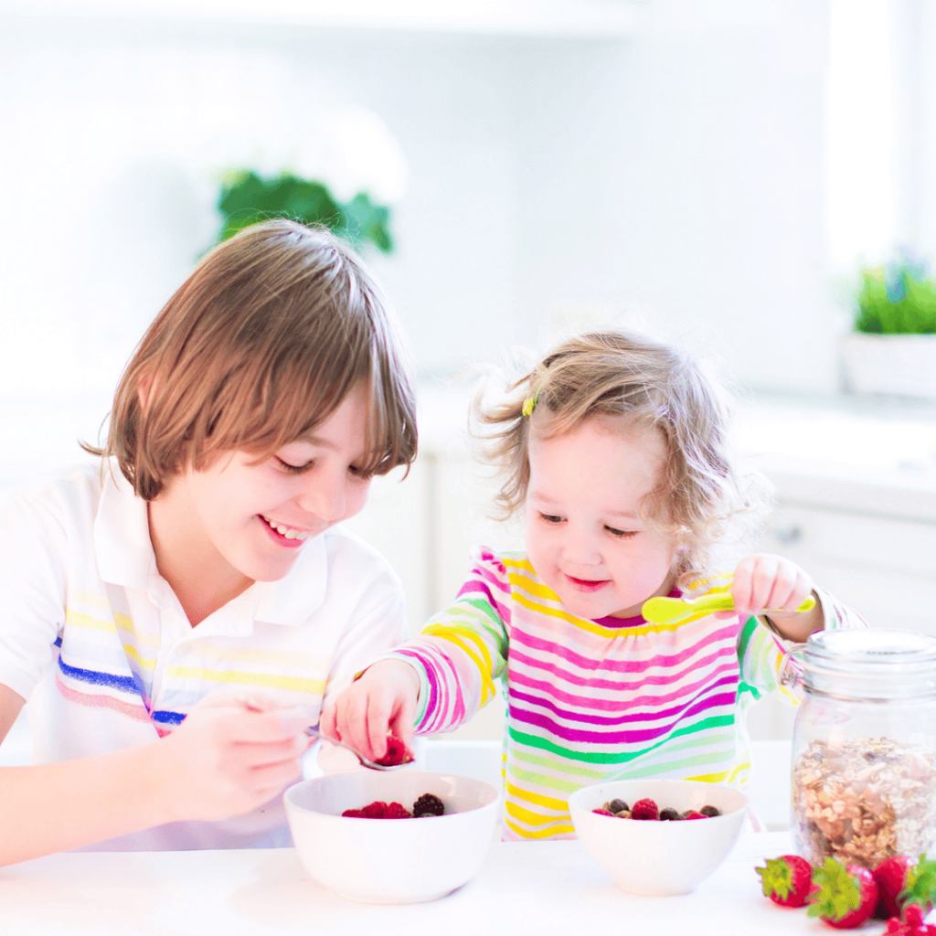 A boy and his toddler sister sit at a kitchen table eating berries. 