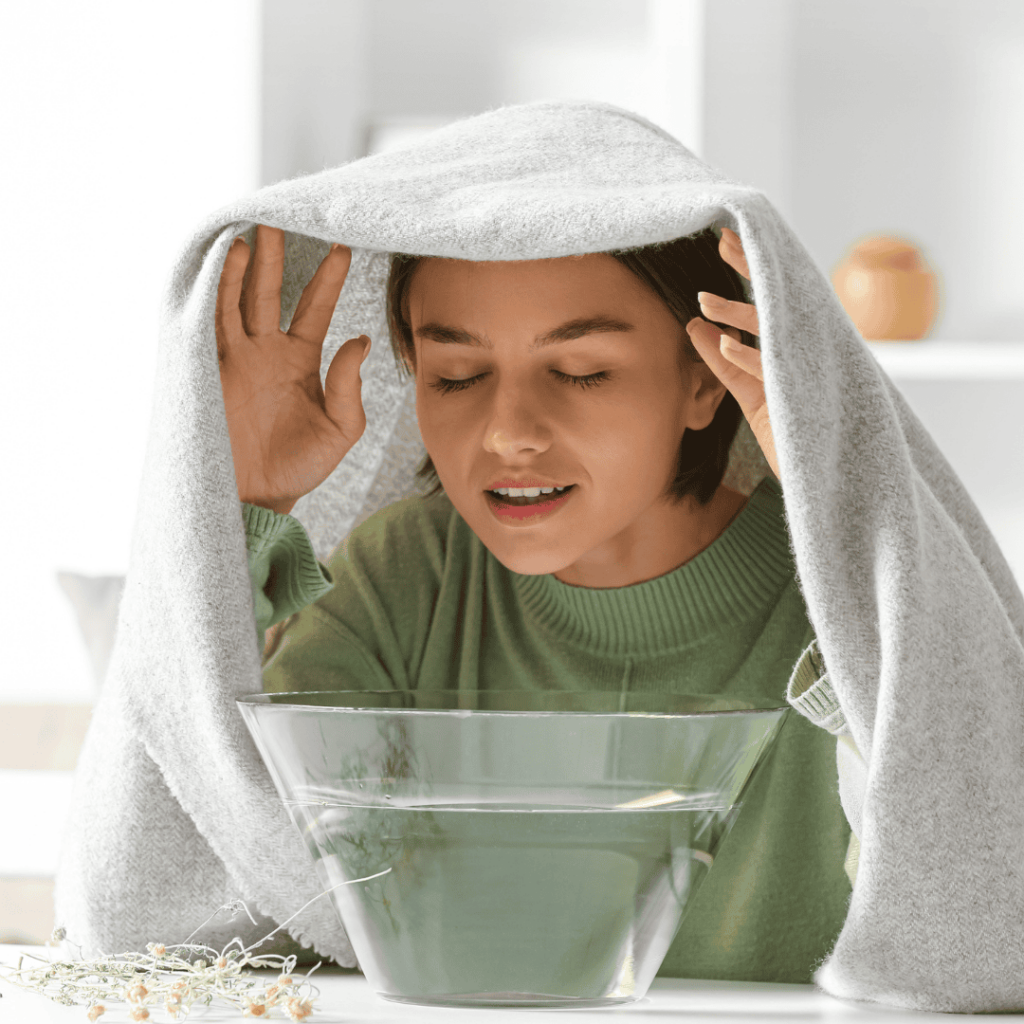 A woman holds a towel over hear head as she inhales steam from a bowel of hot water.
