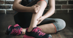 A woman sitting on a gym floor massages the cramp in her calf.