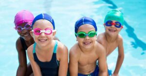 Four children wearing colorful goggles, swim caps, and swim suits pose for a photo in front of a pool.