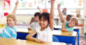 Children sit quietly at desks in a classroom and raise their hands.