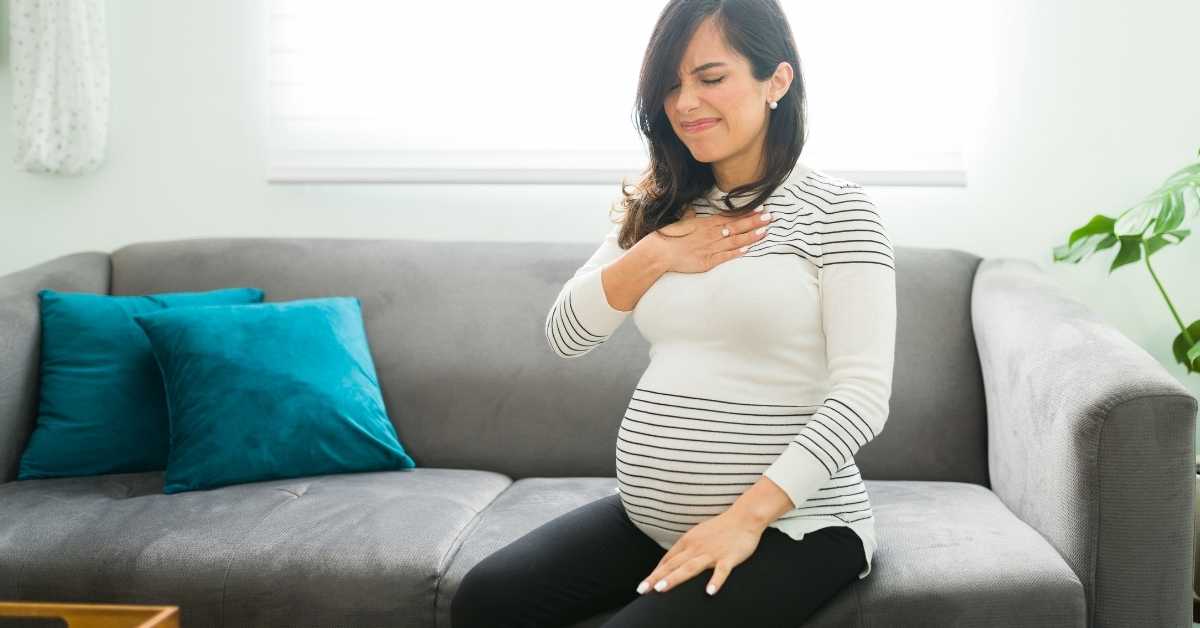 A pregnant woman sits on a couch with her hand clutching her chest and discomfort on her face while experiencing heartburn.