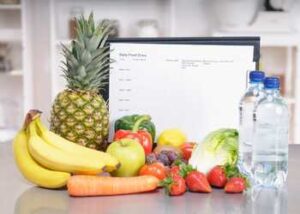 A food diary on a gray kitchen counter surrounded by colorful fruits and vegetables and two water bottles.