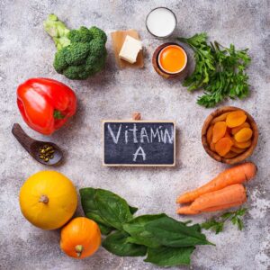 Several foods that are high in Vitamin A lay on a table in a circle, including bell peppers, carrots, broccoli, butter.