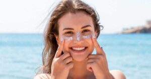 A woman puts sunscreen on her face while at the beach.