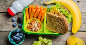 A nutritious lunch of carrots, blueberries, milk, a sandwich on whole grain bread, and a banana packed in a school lunch box.