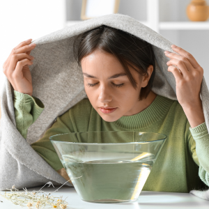 A teenage girl holds a towel over her head while breathing in steam from a bowl of hot water.