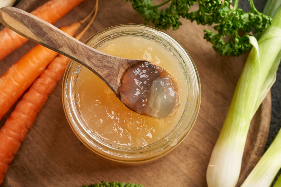 A jar of cooled bone broth rests on a wooden surface surrounded by vegetables.