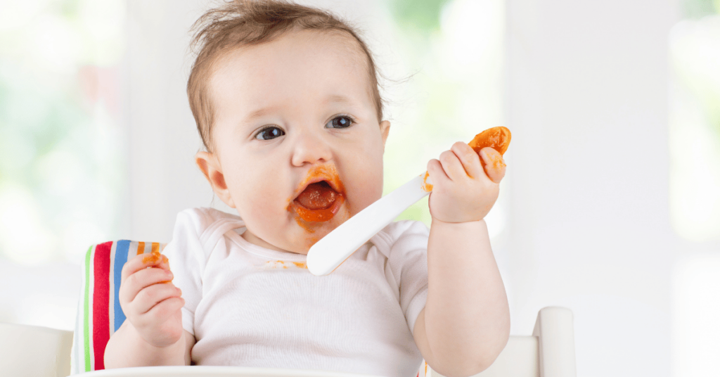 A baby with pureed carrots around her mouth sits in a high chair and holds a spoon.
