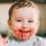 A baby girl has red jam on her hands and face.