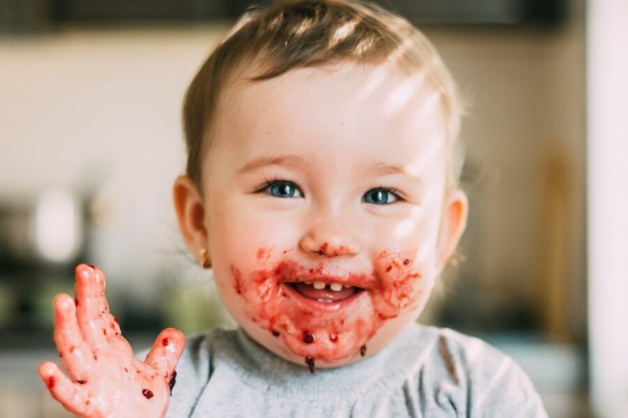 A baby girl has red jam on her hands and face.