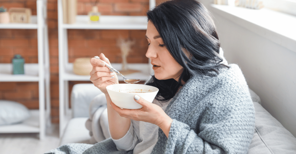 A woman eats a bowl of soup while sitting on a couch.
