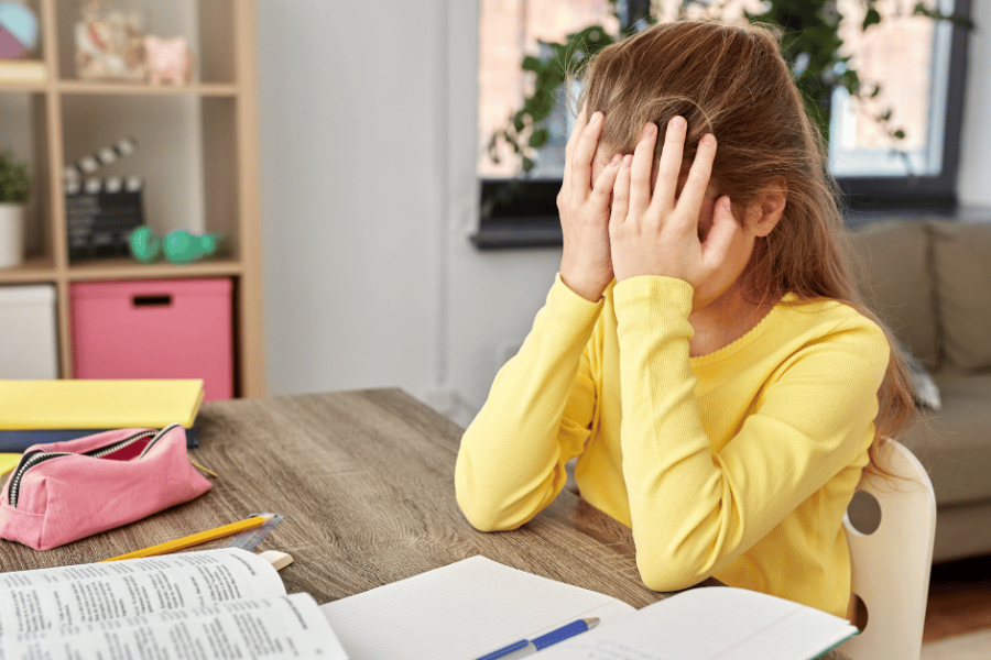 A stressed girl rests her head in her hands while sitting at her desk with her homework in front of her.