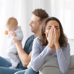 A stressed woman holds her head while her husband plays with their baby in the background.