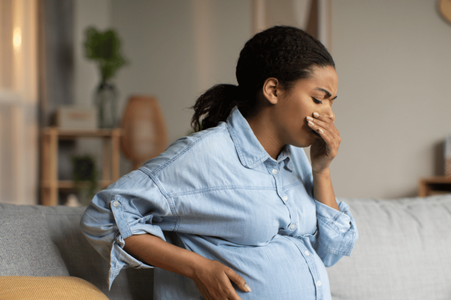 A pregnant woman in a blue shirt sits on a couch looking nauseous.