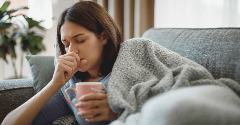 An ill woman lays on her couch while coughing.