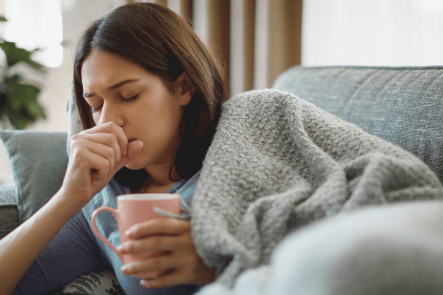 An ill woman lays on her couch while coughing.