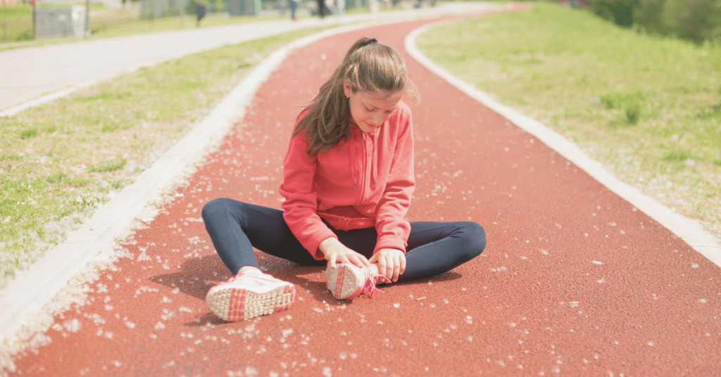 A girl wearing a red sweatshirt sits on the track holding her ankle.