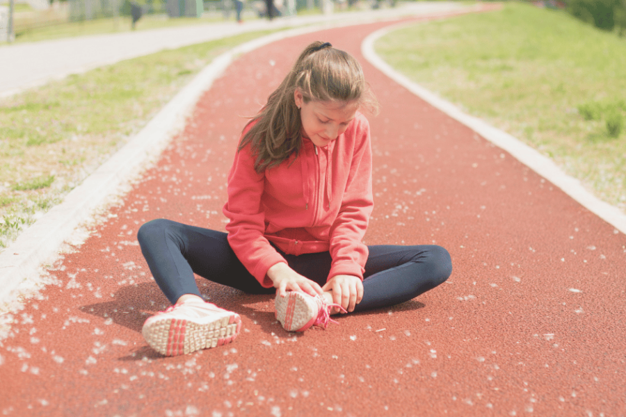 A girl wearing a red sweatshirt sits on the track holding her ankle.