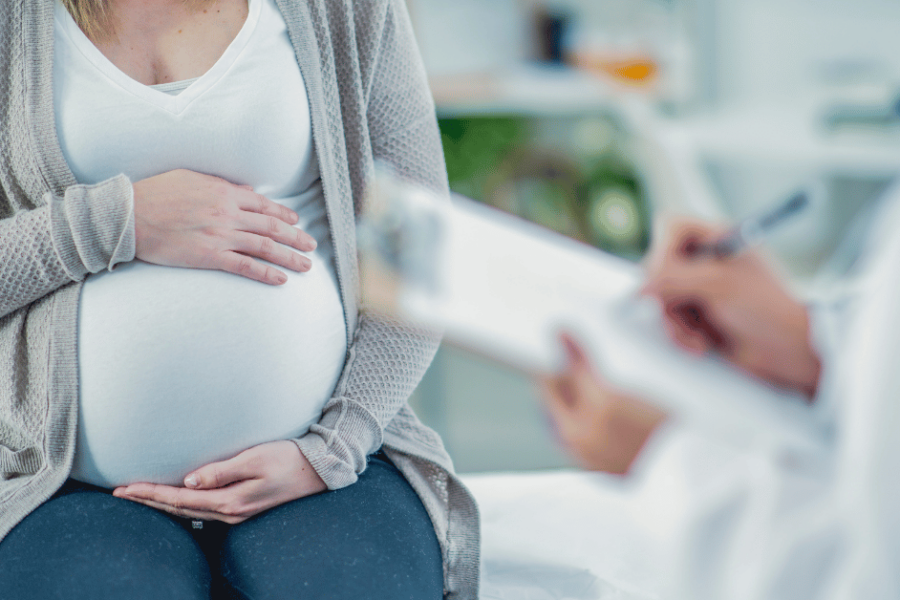 A pregnant woman sits on an exam table while her doctor writes notes on a clipboard.