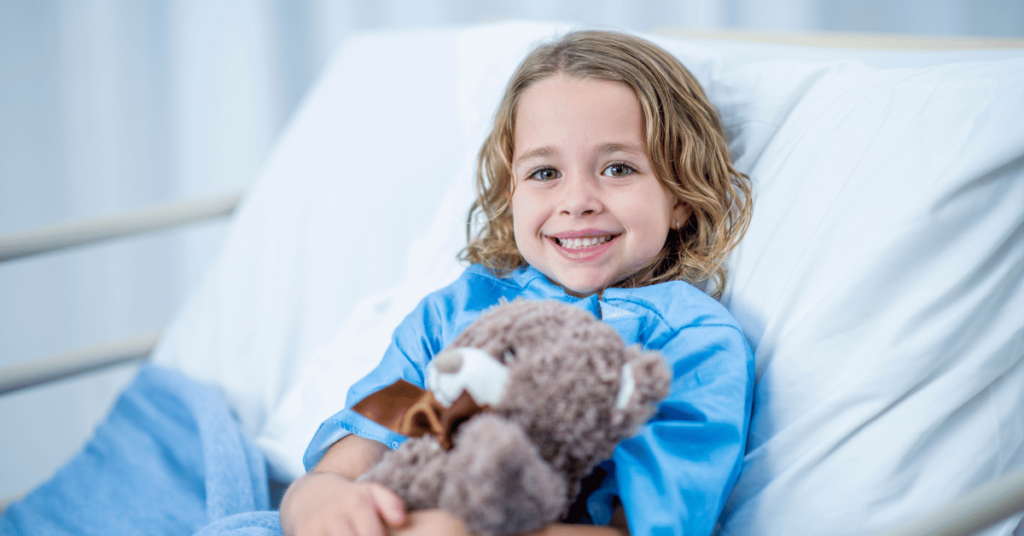 A child in a hospital gown lies in a hospital bed holding a teddy bear.