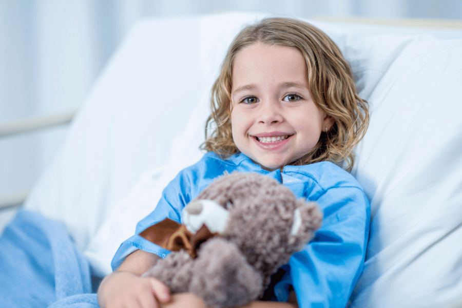 A child in a hospital gown lies in a hospital bed holding a teddy bear.