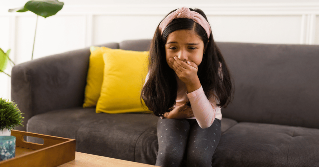 A sick girl sits on the couch holding her stomach and mouth.