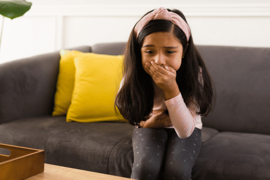 A sick girl sits on the couch holding her stomach and mouth.