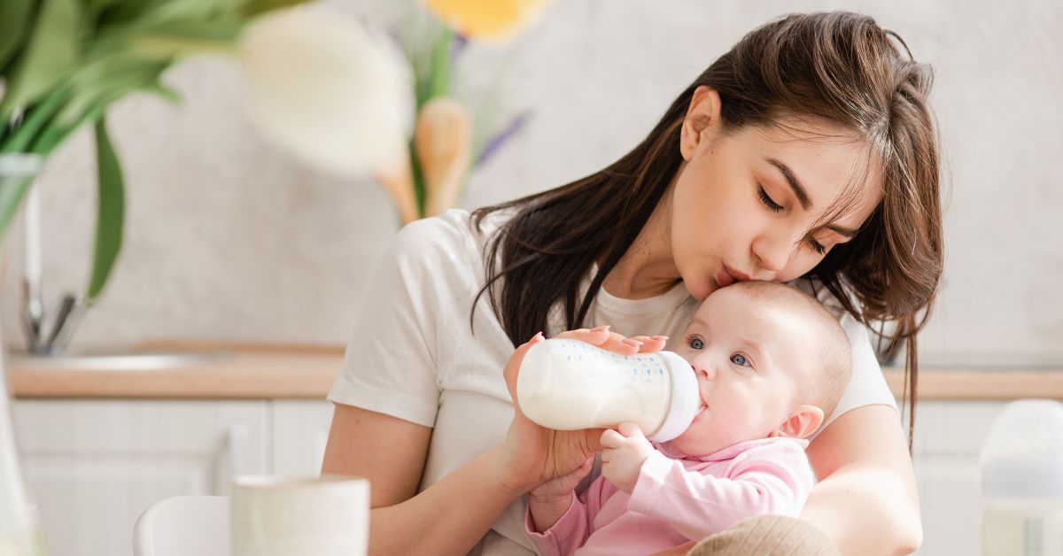 A mom kisses her baby's head while her baby drinks a bottle.