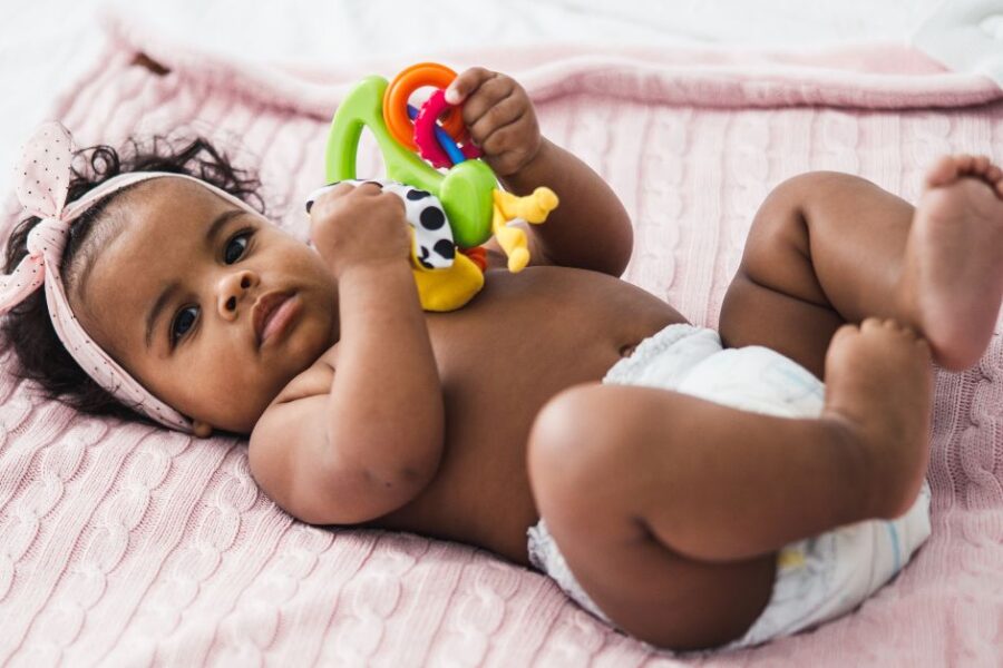 A baby wearing a diaper lays on a pink blanket playing with a toy.