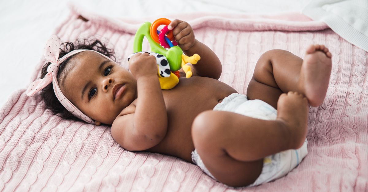 A baby wearing a diaper lays on a pink blanket playing with a toy.
