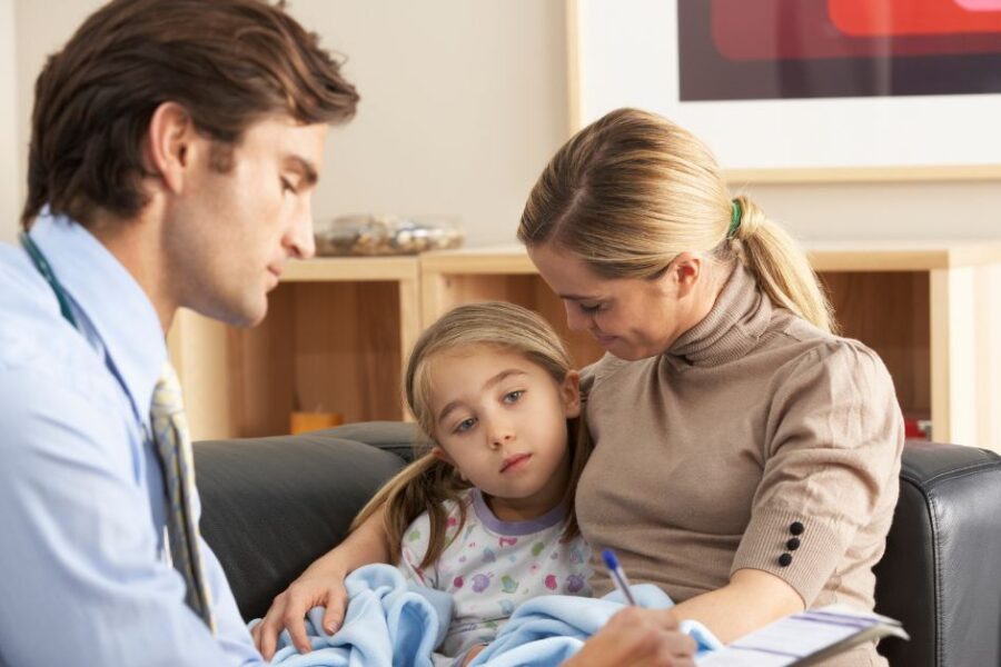 A doctor takes notes while visiting with a sick child and her parent.