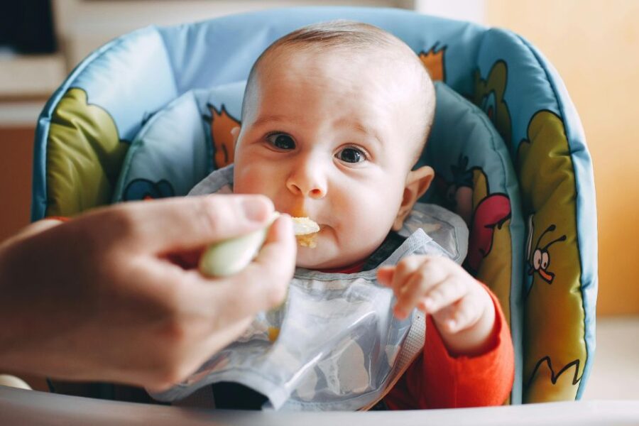 A baby sits in a blue highchair and eats a bite of food from a spoon.