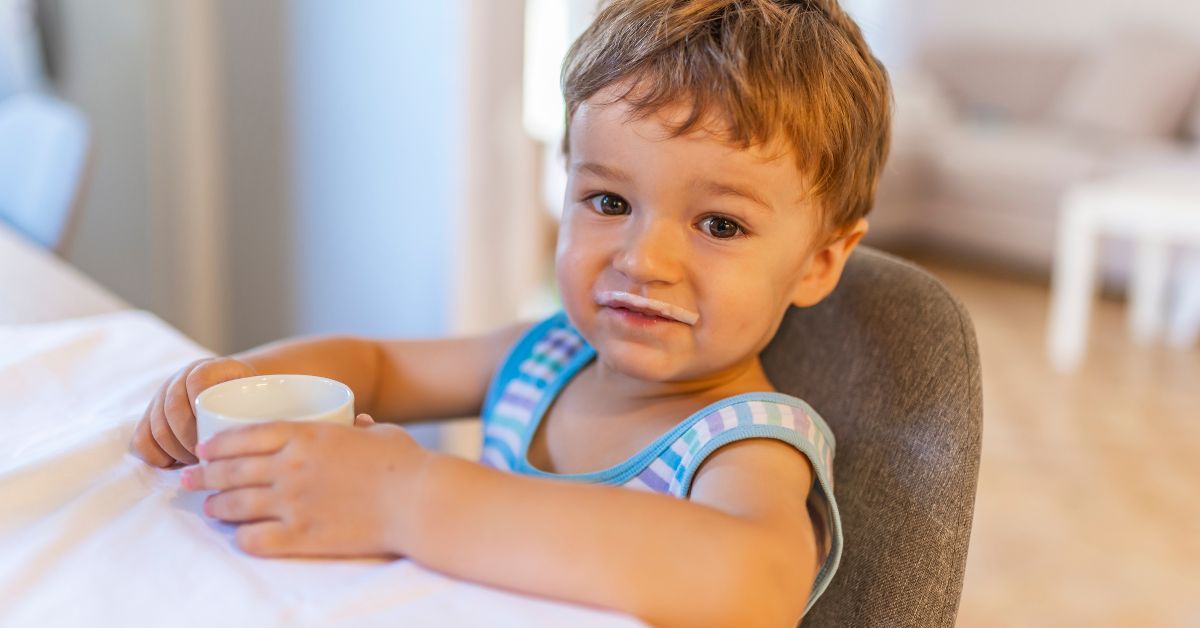 A toddler drinks milk from a small cup while sitting at a table.
