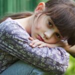 A sullen girl rests her head on her knees while sitting outdoors.